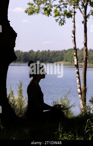 Dieses heitere Bild fängt die Silhouette einer jungen Millennial-Frau ein, während sie Yoga praktiziert und im Herzen der Natur meditiert. Sie sitzt anmutig auf üppig grünem Gras neben einem ruhigen Waldsee und findet innere Ruhe und Verbindung mit der Natur. Diese harmonische Szene ist eine Ode an Achtsamkeit, Ruhe und die heilende Kraft der Natur. Ruhige Yoga Meditation in der Natur. Hochwertige Fotos Stockfoto