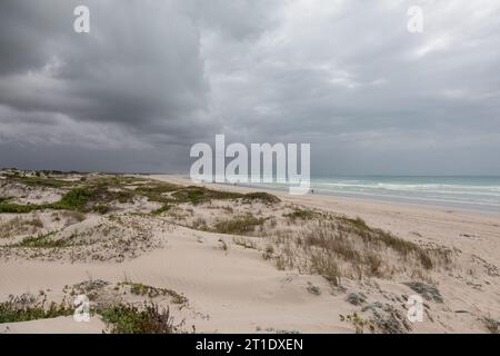 Unter einem grauen und stürmischen Himmel treffen weite Sanddünen auf einen zerklüfteten Strand, an dem ferne Figuren entlang der Küste spazieren Stockfoto