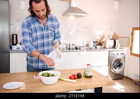 Authentisches Porträt eines gutaussehenden kaukasischen Mannes, Koch, der trockene Gemüse und Kräuter in eine weiße Schüssel legt, während er gesunden Salat in der Küche zubereitete Stockfoto