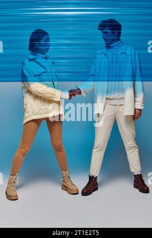 In voller Länge ein stilvolles interrassisches Paar, das Hände hinter blauem Plastik hält, im Studio, Winteroutfit Stockfoto