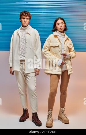 In voller Länge eines interrassischen Paares in warmen Jacken und Lederstiefeln, das sich neben blauem Plastikblech posiert Stockfoto