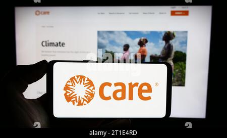 Person mit Handy mit Logo der humanitären Agentur CARE International vor der Webseite. Schwerpunkt auf dem Display des Telefons. Stockfoto