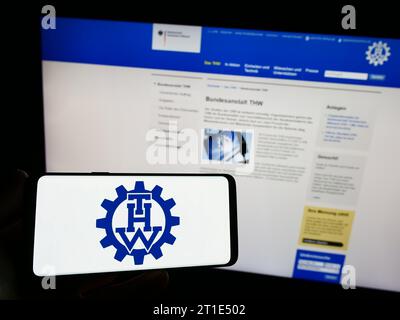 Person mit Smartphone mit Logo der Bundesanstalt Technisches Hilfswerk (THW) vor der Website. Schwerpunkt auf dem Display des Telefons. Stockfoto