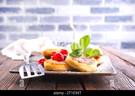 Traditionelle süße Syrniki- oder Hüttenkäsepfannkuchen serviert mit süßer Sahne und frischen Himbeeren auf einem weißen Teller über dem Tisch. Stockfoto