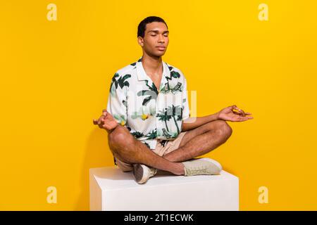 Ganzkörperfoto des attraktiven jungen Mannes weiße Plattform Meditation gekleidet stilvolle Palmen drucken Kleidung isoliert auf gelbem farbigen Hintergrund Stockfoto