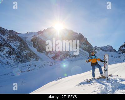 Ein Mann mit Helm und Snowboard steht vor dem Abstieg vom Berg, vor der Kulisse epischer Felsen mit aufgehender Sonne. Snowboarder Stockfoto