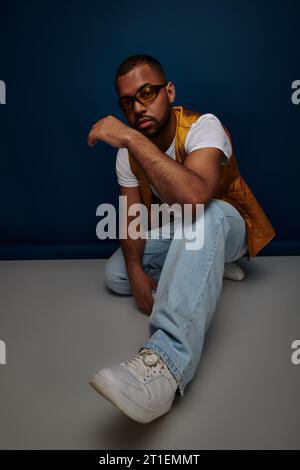 Hübscher junger Mann in Weste und Jeans, der vor dunkelblauem Hintergrund hockt und posiert, Modekonzept Stockfoto