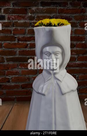 Vertikales Innenbild mit einer weißen porzellanvase in Form einer Nonne, mit gelben Blüten darin, auf einem Holzboden, vor einer natürlichen Ziegelwand Stockfoto