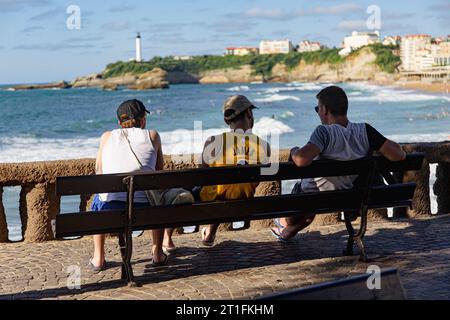 Drei Männer sitzen auf einer Bank und starren auf den Strand in Biarritz. Leuchtturm weit weg im Hintergrund. Stockfoto