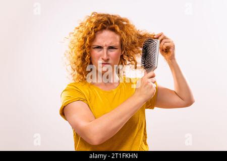 Wütende Ingwerfrau, die Haarbürste hält und ihr lockiges Haar kämmt. Stockfoto