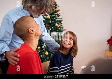 Großvater steht lächelnd und plaudert mit Tochter und Enkelkind, die am Weihnachtstisch sitzen. Stockfoto