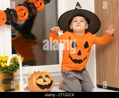 Halloween-Konzept. Ein kleiner, fröhlicher, gutaussehender Junge mit Zaubererhut und orangefarbenem Pullover sitzt auf einem Tisch in der Küche vor dem Hintergrund eines Stockfoto