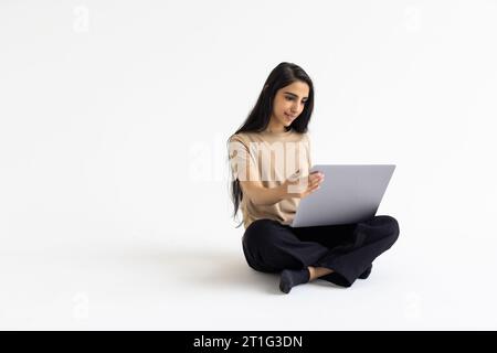 Online-Assistent, Chatbot, künstliche Intelligenz im Alltag. Lächelnde, schöne junge indianerin in lässigem Outfit mit Laptop auf weißem Wal Stockfoto