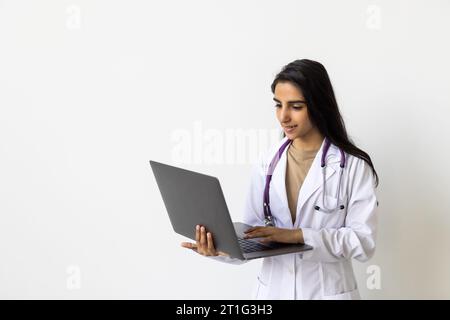 Fröhliche junge attraktive hinduistische Ärztin in Uniform arbeitet am Computer isoliert auf weißem Hintergrund. Soziale Distanz während covid-19-Pandem Stockfoto