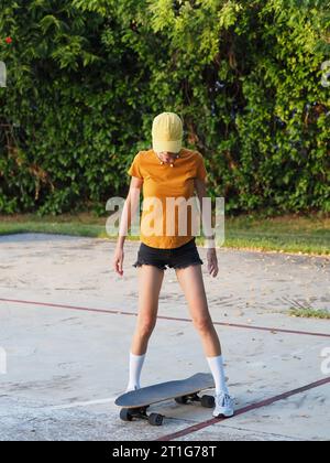 Frau, die zum ersten Mal auf Skateboard schaut, mit farbenfroher gelber Mütze und Shorts. Stockfoto