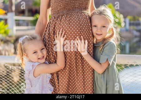 Die Liebe der Schwestern blüht, während sie den schwangeren Bauch ihrer Mutter zärtlich umarmen und ihre Vorfreude und Zuneigung für ihre bald ankommenden Geschwister teilen Stockfoto