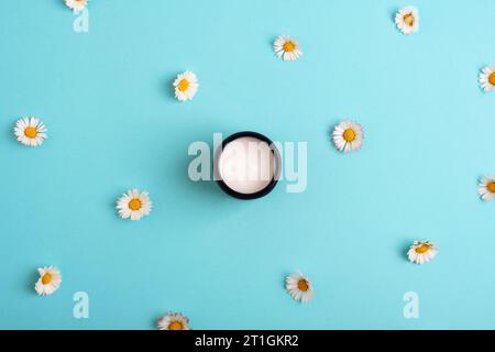 Kosmetische Creme in einem Glas und Gänseblümchen auf blauem Hintergrund. Draufsicht, flach. Stockfoto