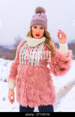 Porträt eines jungen blonden Models mit rosa Pelzjacke, Winterstiefeln und lila Hut im Schnee. Lifestyle Stockfoto