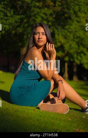 Eine hübsche junge, brünette Latina mit langen, glatten Haaren und einem engen grünen Kleid. Porträt eines Mädchens, das auf dem Gras sitzt, in einem Park mit Bäumen drin Stockfoto