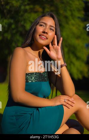 Eine hübsche junge, brünette Latina mit langen, glatten Haaren und einem engen grünen Kleid. Porträt eines jungen Mädchens, das in einem Park träumt und sich Dinge vorstellt Stockfoto