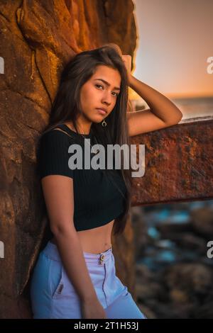 Eine junge hübsche Brünette Latina mit langen glatten Haaren in einem kurzen schwarzen T-Shirt und rosa Hose. Porträt der Frau, die sich auf eine Skulptur mit Schorf stützt Stockfoto