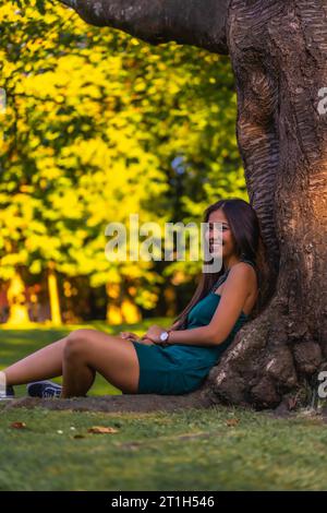 Eine junge hübsche Latina Brünette mit langen, glatten Haaren, die sich in einem grünen Kleid an einen Baum lehnt. Am Baum im Park sitzen Stockfoto