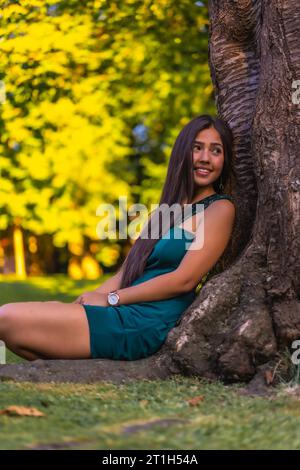 Eine junge hübsche Brünette Latina mit langen, glatten Haaren, die sich in einem grünen Kleid an einen Baum lehnt. Auf dem Gras neben einem Baum im sitzen Stockfoto