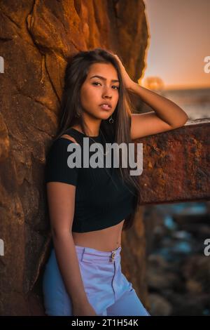 Eine junge hübsche Brünette Latina mit langen glatten Haaren in einem kurzen schwarzen T-Shirt und rosa Hose. Porträt der Frau, die sich auf eine Skulptur mit Schorf stützt Stockfoto