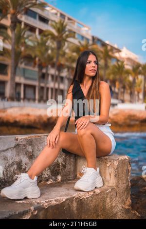 Sommerleben, junge brünette Kaukasier an der Mittelmeerküste, die auf einem Felsen am Meer sitzt Stockfoto