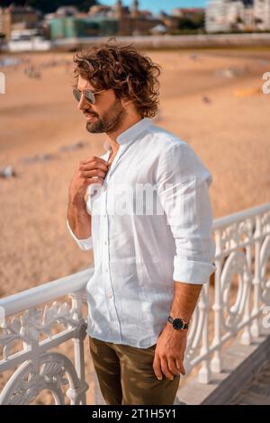 Sommer-Lifestyle mit einem jungen dunkelhaarigen Kaukasier mit langem Bart, Sonnenbrille und weißem Hemd am Strand. San Sebastian, Baskenland. Stockfoto