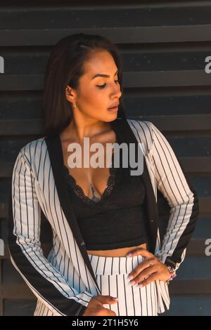Lifestyle, glatthaariges lateinamerikanisches Brünette-Mädchen in weißem Anzug und schwarzen Streifen. Schwarzer Hintergrund. Sieht sexuell anders aus, sieht anders aus Stockfoto