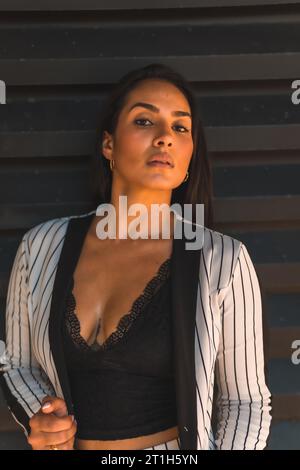Lifestyle, glatthaariges lateinamerikanisches Brünette-Mädchen in weißem Anzug und schwarzen Streifen. Schwarzer Hintergrund. Sieht sexuell anders aus, sieht anders aus Stockfoto