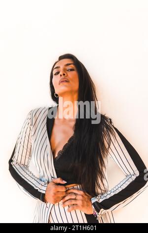 Lifestyle, glatthaariges lateinamerikanisches Brünette-Mädchen in weißem Anzug und schwarzen Streifen. Weißer Hintergrund. Sieht sexuell anders aus, sieht anders aus Stockfoto