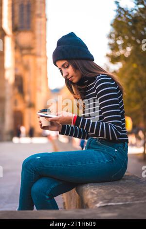 Lifestyle, brünette Kaukasierin, die eine Nachricht mit ihrem Handy in der Stadt schickt. Entspannen Sie sich bei einem Kaffee zum Mitnehmen neben einer wunderschönen Kirche im Stockfoto