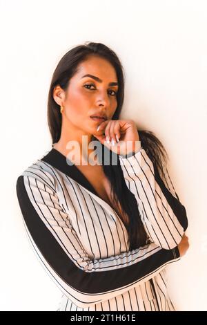 Lifestyle, glatthaariges lateinamerikanisches Brünette-Mädchen in weißem Anzug und schwarzen Streifen. Weißer Hintergrund. Sieht sexuell anders aus, sieht anders aus Stockfoto