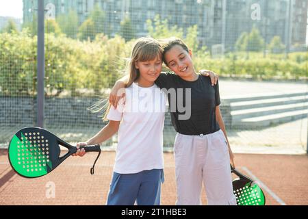 Kinder- und Sportkonzept. Porträt von lächelnden Mädchen, die auf dem Padel-Platz mit Schlägern und Tennisbällen im Freien posieren Stockfoto