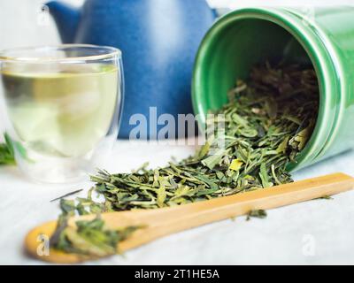 Stillleben von grünem Tee Long jing Drachenbrunnen aus China. Teeblätter auf Leinentischtuch mit grünem Tee in Tasse, Teekanne und Bambuslöffel. Stockfoto