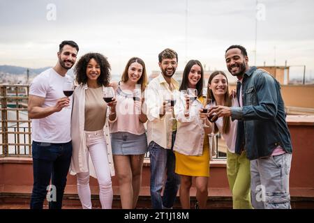 Vielfältige Gruppe von Freunden bei Drinks und Toasten auf einer Dachparty. Multirassische junge, glückliche Gruppe von Millennials, die Wein auf einer Terrasse trinken Stockfoto