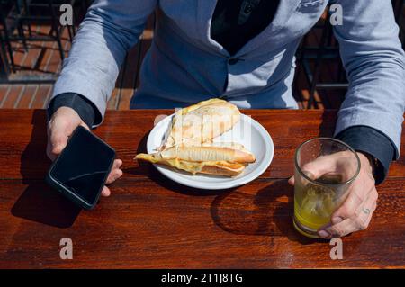 Blick von oben auf die kaukasischen Hände eines nicht erkennbaren Geschäftsmannes, der vor dem Restaurant sitzt und ein Frühstück mit Schinken-Käse-Sandwich mit Orangensaft A genießt Stockfoto