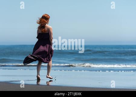 Rückansicht der rothaarigen Frau in dunkelviolettem langem Kleid, die auf Sandstrand in Richtung Wasser auf dem Hintergrund des blauen Meeres an sonnigem Tag mit klarem Himmel geht. Fu Stockfoto