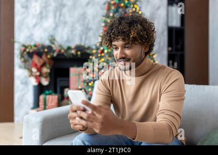 Junger gutaussehender hispanischer Mann zu Hause auf Sofa im Wohnzimmer für Weihnachten in der Nähe von Baum, Mann mit Telefon glücklich lächelnd Tippen Online-Textnachricht und Surfen Internet-Seiten. Stockfoto