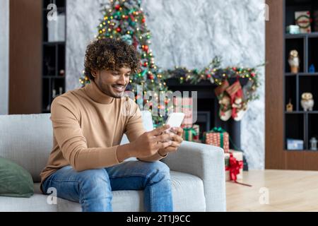 Junger gutaussehender hispanischer Mann zu Hause auf Sofa im Wohnzimmer für Weihnachten in der Nähe von Baum, Mann mit Telefon glücklich lächelnd Tippen Online-Textnachricht und Surfen Internet-Seiten. Stockfoto