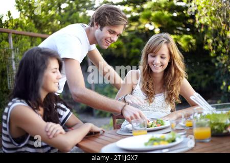 Glücklich, Freunde und Essen servieren im Hinterhof mit Salat an einem Tisch mit hungrigen Leuten und Lächeln. Mittagessen, Garten und Gruppe bereit zum Essen mit Stockfoto