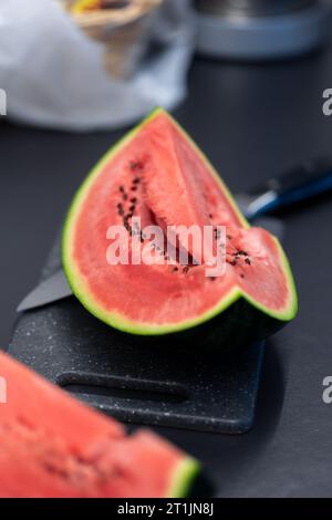 Vertikales Porträt eines Viertelstücks einer grün geschnittenen Wassermelone, das auf einem Schneidebrett aus Kunststoff neben einem Messer liegt. Die Scheibe der Frucht ist bereit Stockfoto