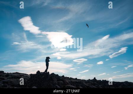 Silhouette eines Fotografen, der mit Kamera steht und ein Foto auf dem Berg macht, und eines Vogels, der im blauen Himmel fliegt Stockfoto