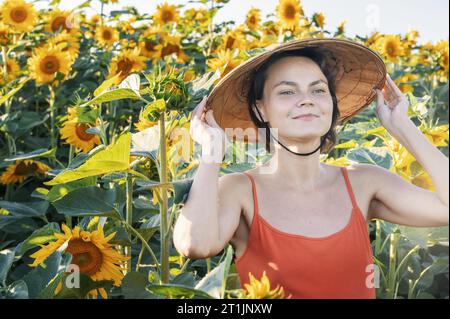 Schöne kaukasische Frau in Sonnenblumen, Porträt einer Frau mit vietnamesischem Hut, die bei Sonnenuntergang durch ein blühendes Sonnenblumenfeld geht. Frieden, Stockfoto