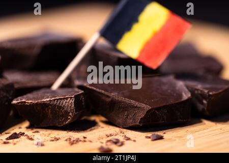 Ein Porträt einiger grober geschnittener Stücke köstlicher brauner, dunkler belgischer Schokolade, die auf einer hölzernen Oberfläche liegen, mit einer kleinen verschwommenen belgischen Flagge in b Stockfoto