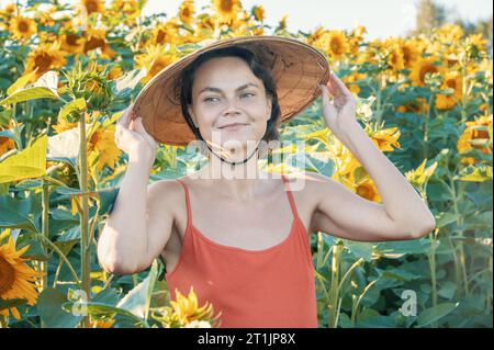 Schöne kaukasische Frau in Sonnenblumen, Porträt einer Frau mit vietnamesischem Hut, die bei Sonnenuntergang durch ein blühendes Sonnenblumenfeld geht. Frieden, Stockfoto