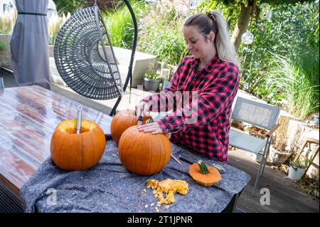 Blonde Frau, die die Spitze eines Kürbis öffnet, um halloween-Dekorationen mit einem Messer zu schnitzen Stockfoto