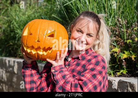 Die blonde Frau im Garten präsentiert zu Halloween einen selbst geschnitzten Kürbis mit Gesicht Stockfoto